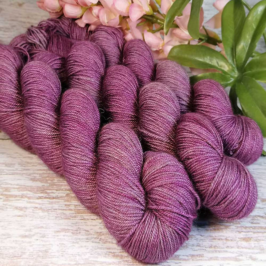 Handgefertigte Mesiya Wolle in sanftem Mauve, vier dicke Stränge auf Holzuntergrund mit grünen Blättern und rosa Blüten im.