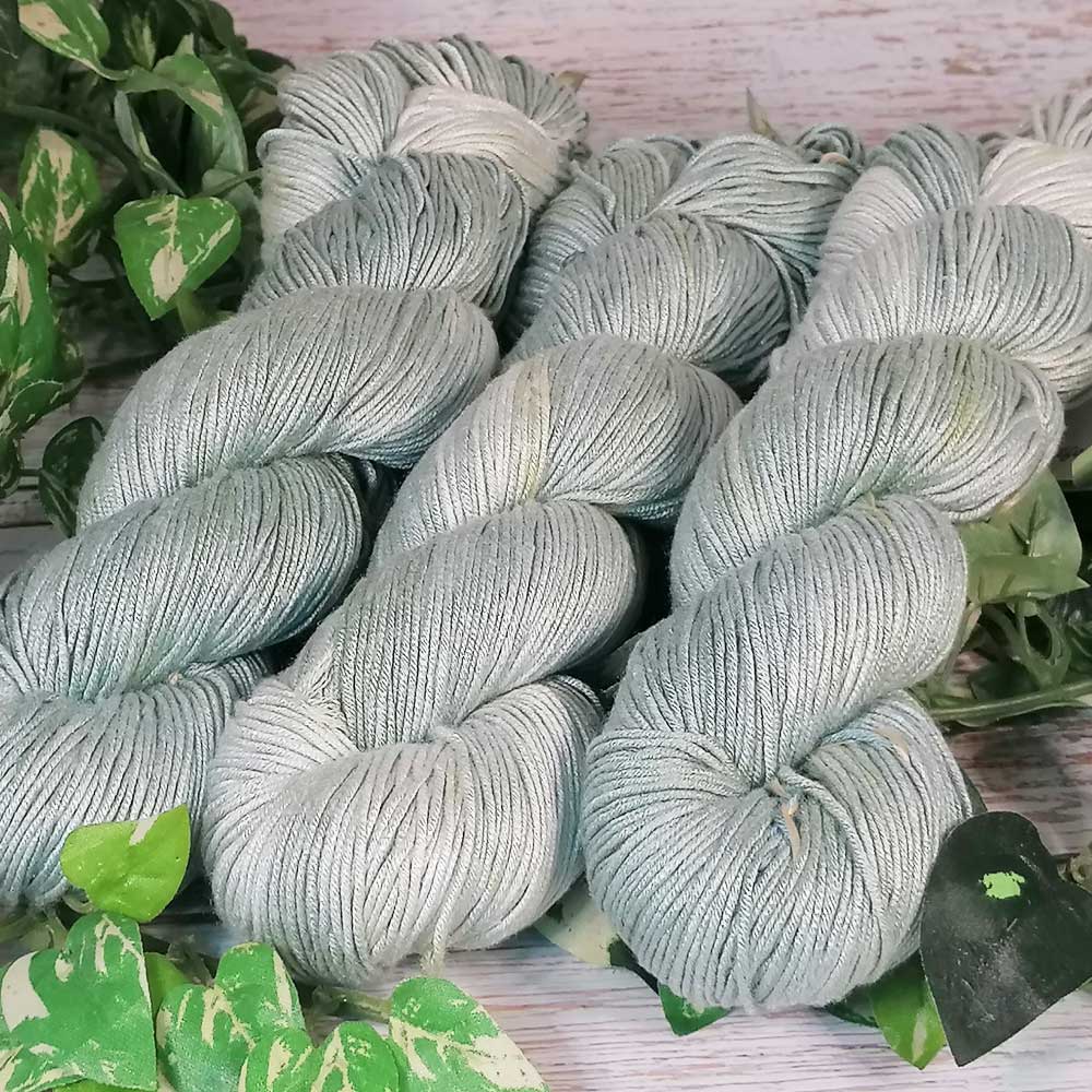 Drei handgefertigte, hellgraue Garnstränge mit feiner Textur, liegend auf hellem Holz mit grünen Blättern im Hintergrund.
