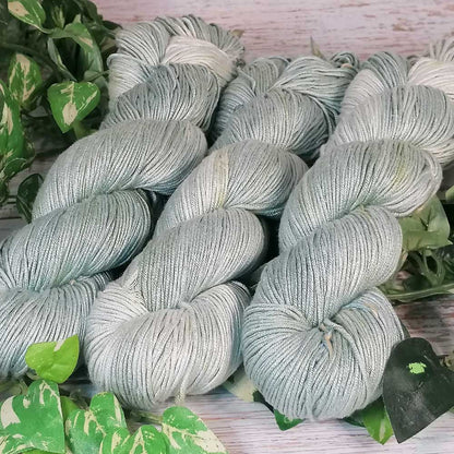 Drei handgefertigte, hellgraue Garnstränge mit feiner Textur, liegend auf hellem Holz mit grünen Blättern im Hintergrund.