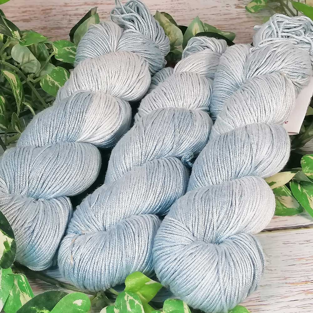 Handgefertigte, hellblaue Arcadia-Garne mit weicher Textur, auf hellem Holz mit grünen Blättern arrangiert.