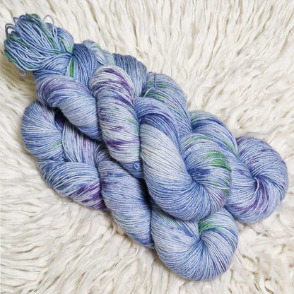 Drei Stränge hellblau-lila melierte Wolle mit grünen und blauen Sprenkeln auf weißem Fellhintergrund.