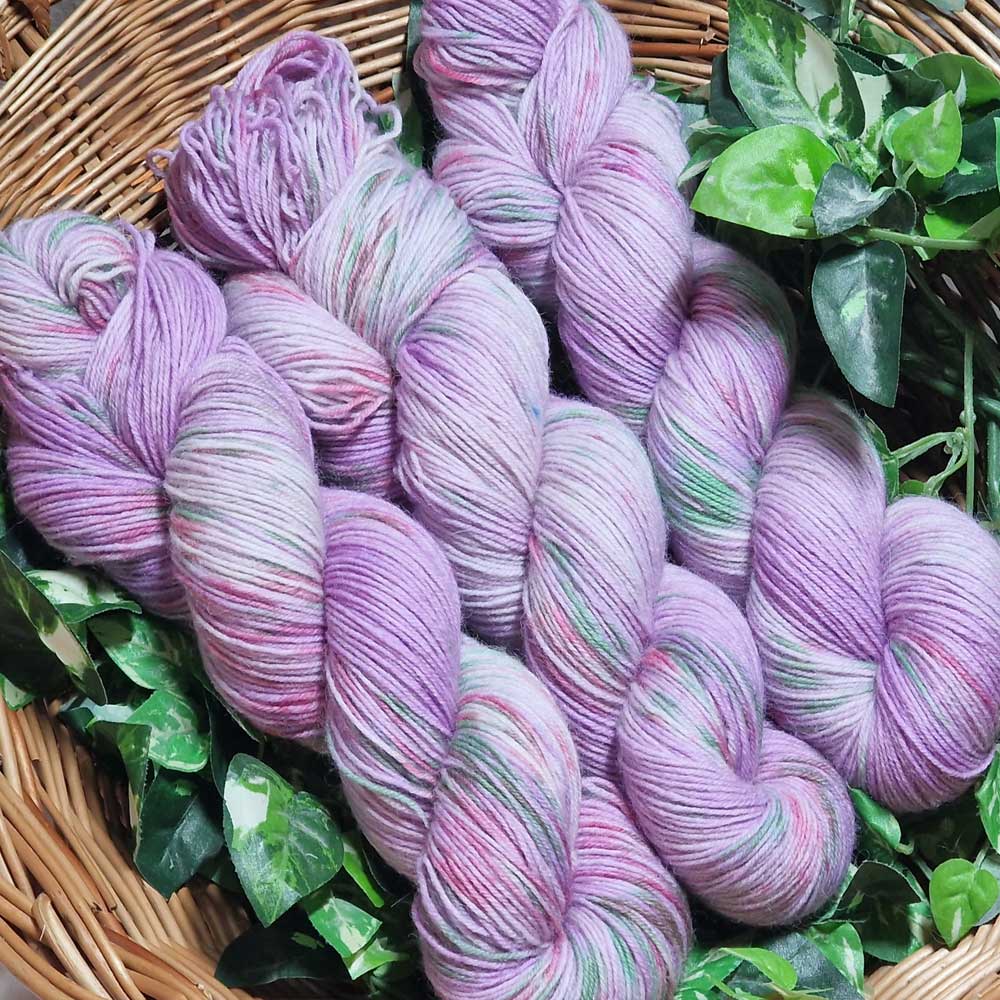 Mehrere Stränge rosa-lila gemustertes Garn in einem Weidenkorb mit grünen Blättern.