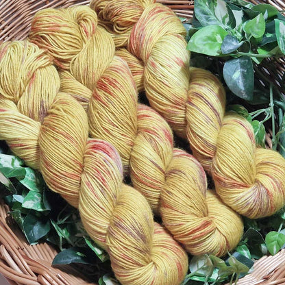 Gestricktes Garn in warmem Senfgelb mit roten Sprenkeln in Flechtkorb auf grünem Blätterhintergrund.