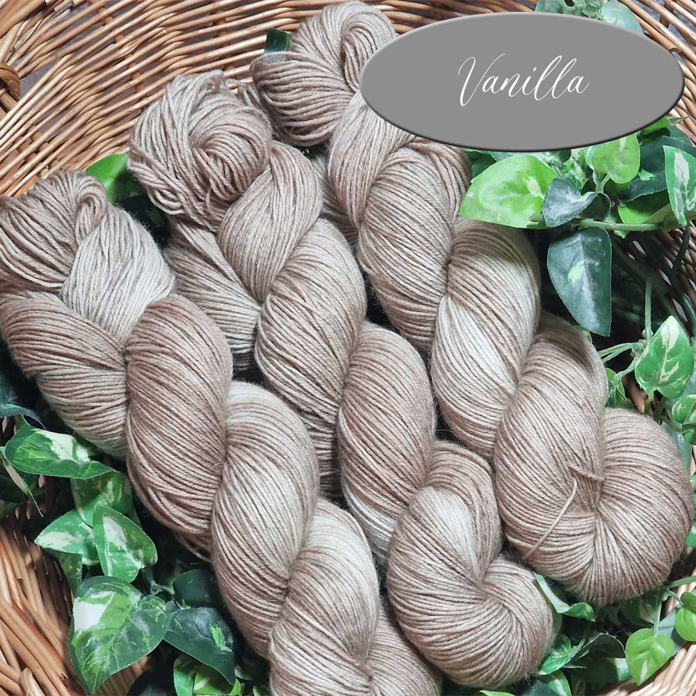 Mehrere helle, vanillefarbene Sockengarne in Strängen, locker drapiert mit grünen Blättern im Hintergrund.