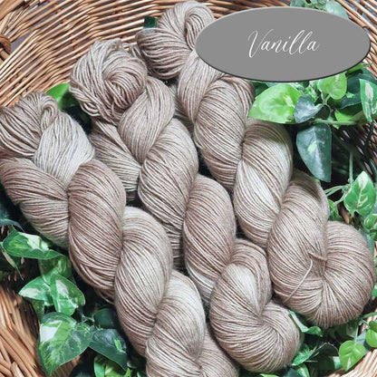 Mehrere helle, vanillefarbene Sockengarne in Strängen, locker drapiert mit grünen Blättern im Hintergrund.