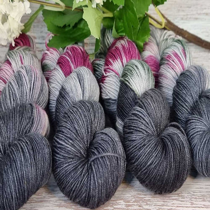 Handgefertigte Garnstränge in dunklem Anthrazitgrau mit Akzenten in Pink, Weiß und Grün, locker auf hellem Holz arrangiert.