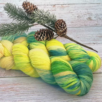 Handgefertigte Wollstränge in Gelb-, Grün- und Blautönen mit Nadelzweig und Kiefernzapfen auf hellem Holzhintergrund.