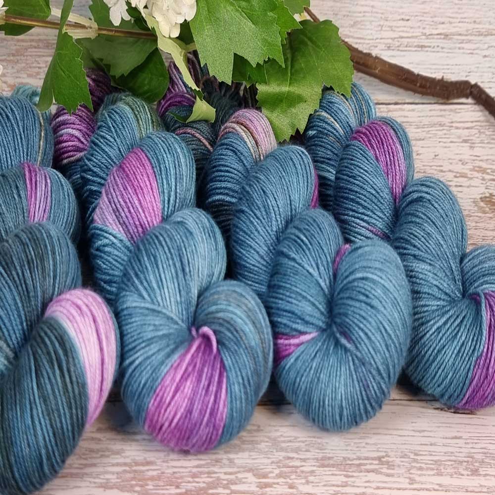 Handgefertigte Wollstränge in Blau mit Akzenten in Lila und Pink, dicht nebeneinander auf hellem Holztisch liegend, umgeben.