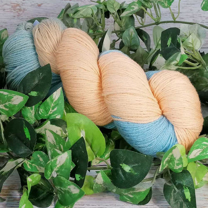 Handgefertigtes Merino Baby Garn in sanften Pastelltönen Beach und Lagune, umgeben von grünen Blättern auf Holzuntergrund.