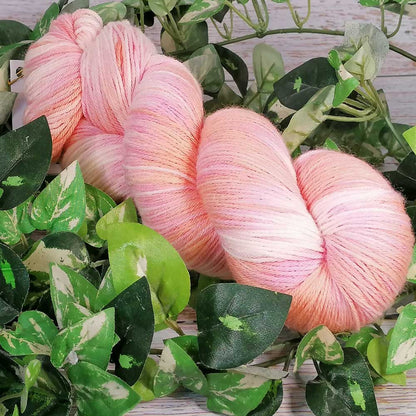 Handgefertigtes Merino Baby Garn in sanften Rosa- und Pfirsichtönen, umgeben von grünen Blättern.