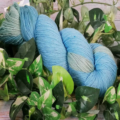 Handgefertigtes Strickgarn in sanften Blau- und Grautönen, umgeben von grünen Blättern, weiches Merino Baby Garn.