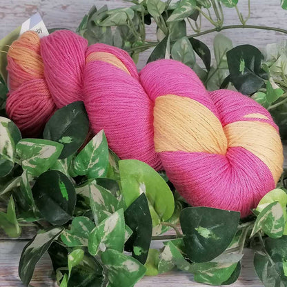 Handgefertigtes Merino Baby Garn in leuchtendem Pink und warmem Gelb, liegt dekorativ zwischen grünen Blättern auf hellem.