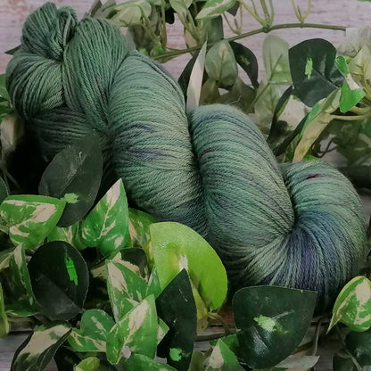 Handgefertigtes Strickgarn in Grüntönen, sanft meliert, umgeben von grünen Blättern, natürliche Merino Qualität.