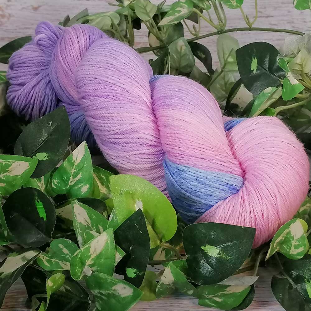 Handgefertigtes Merino-Garn in sanften Pastelltönen von Rosa, Blau und Lila, umgeben von grünen Blättern.